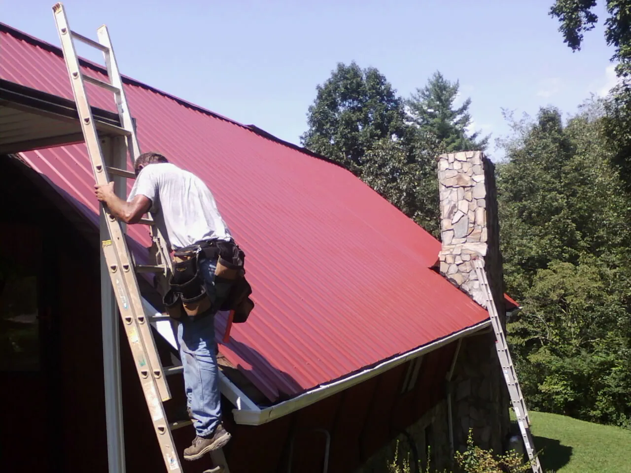 Roofer on a ladder beside a red metal roof and stone chimney — Soffit & Fascia Repair by Roofing Brothers in Harrison